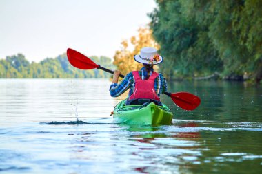 Woman in elegant straw hat canotier paddling green kayak at Danube river at summer. Concept of recreation and adventure