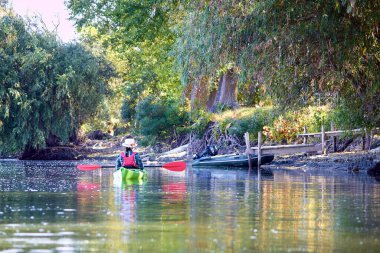 Zarif hasır şapkalı kadın Tuna nehrinin kıyısındaki yeşil kanoda oturuyor..
