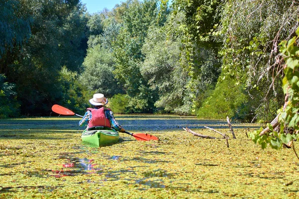 Woman in elegant canotier paddling green kayak in wilderness river overgrown duckweed near thickets of trees and wildgrapes at sunny summer day. Concept of recreation and adventure