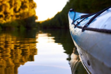 Bow (prow) of blue kayak against a background of green summer trees illuminated by the rays of the setting sun at the shore of Danube river. Kayaking on peaceful calm lake or river