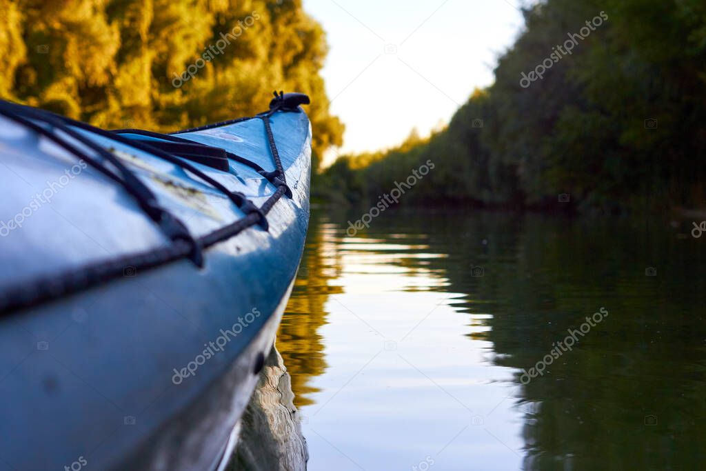 Blue kayak against a background of green summer trees illuminated by the rays of the setting sun at the shore of Danube river. Kayaking on peaceful calm lake or river