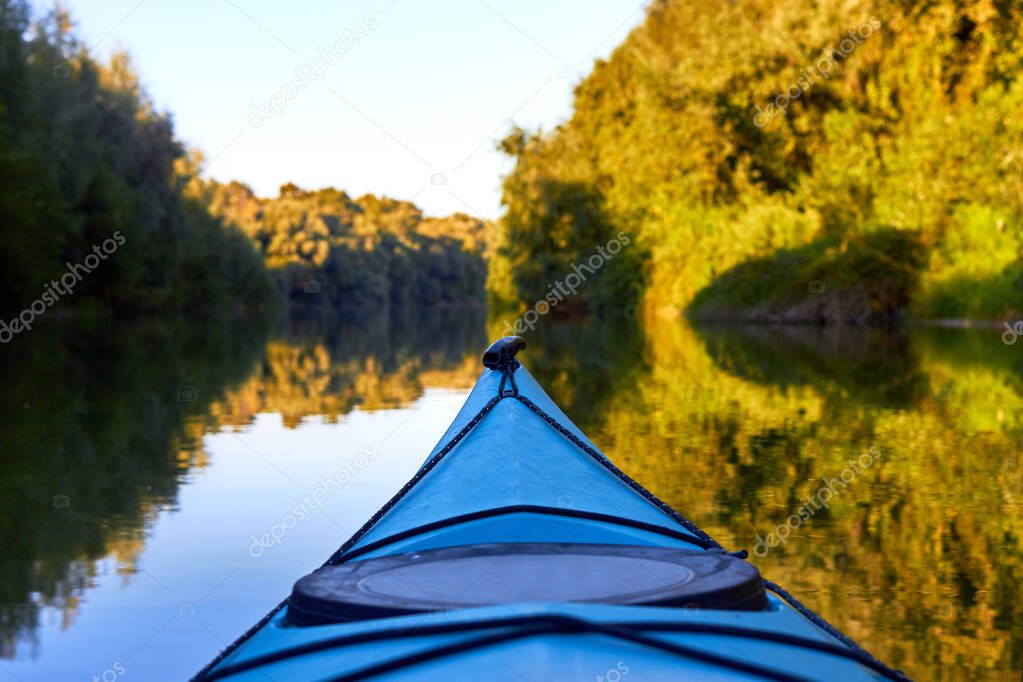Bow (prow) of blue kayak against a background of green summer trees illuminated by the rays of the setting sun at the shore of Danube river. Kayaking on peaceful calm lake or river