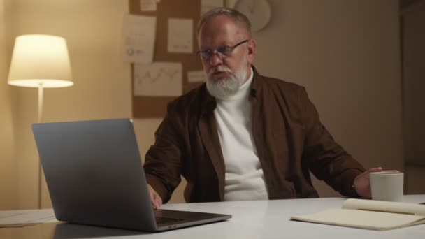 一位老年人晚上在笔记本电脑前工作 喝咖啡 — 图库视频影像