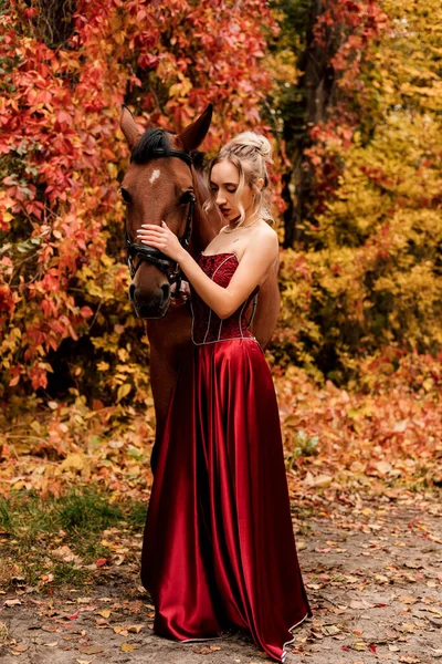 Jeune belle femme en robe bordeaux se tient près d'un cheval dans la forêt d'automne — Photo