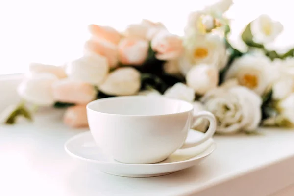 Uma xícara branca e um disco ficam perto das flores em um fundo claro — Fotografia de Stock