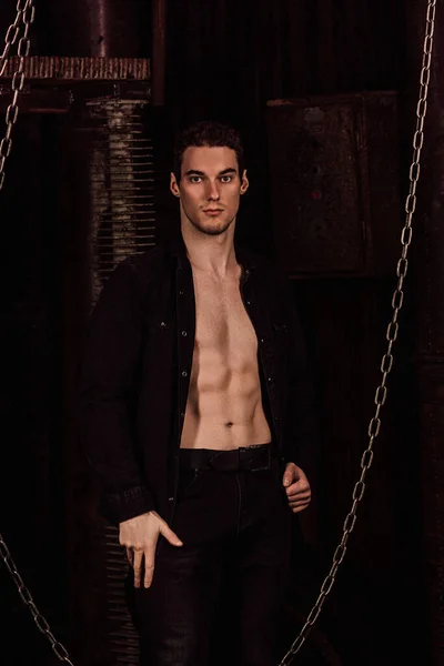 一个穿着衬衫的帅哥站在一间黑暗的房间里，旁边有铁链和管子 — 图库照片