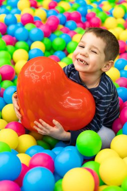 Küçük bir çocuk parlak topların içinde oturuyor ve elinde kırmızı bir top tutuyor. 