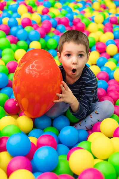 Küçük bir çocuk parlak topların içinde oturuyor ve elinde kırmızı bir top tutuyor. 