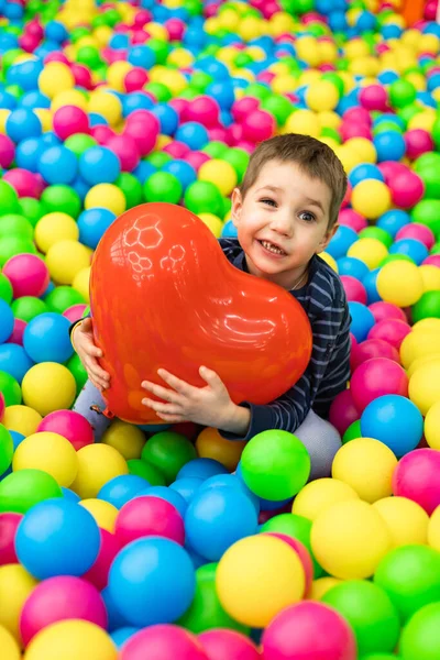 Küçük bir çocuk parlak topların içinde oturuyor ve elinde kırmızı bir top tutuyor. — Stok fotoğraf