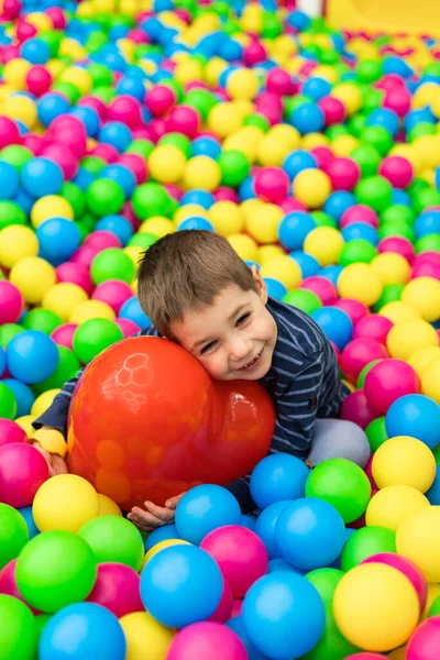 Küçük bir çocuk parlak topların içinde oturuyor ve elinde kırmızı bir top tutuyor. 