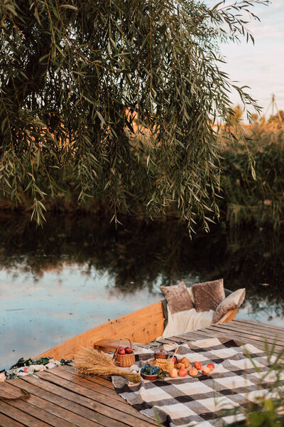On a plaid blanket are fruits and sweets on the pier by the water