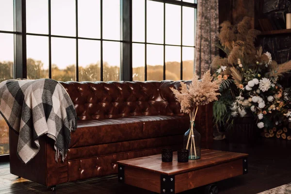 Un vaso con fiori secchi poggia su un tavolo di legno accanto al divano e alla finestra. — Foto Stock