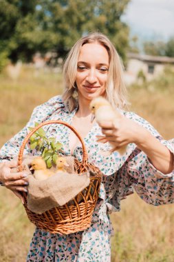 Bir kadın bir elinde ördek yavrusu, diğer elinde ördek yavrusu olan bir sepet tutar.