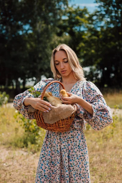 Uzun elbiseli genç bir kadın bir sepet ördek yavrusu tutuyor ve gülümsüyor.