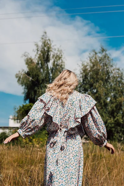 Une fille aux cheveux blonds dans une longue robe se tient avec son dos dans un champ — Photo