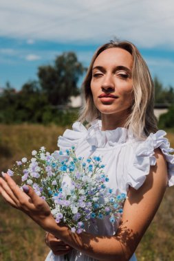 Genç ve güzel bir kız elinde çiçeklerle gülümsüyor.