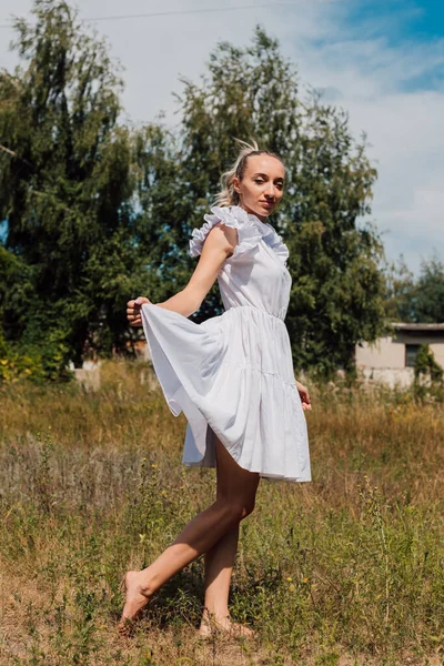 Açık renk elbiseli genç bir kız bir tarlada duruyor ve eliyle bir elbise tutuyor.