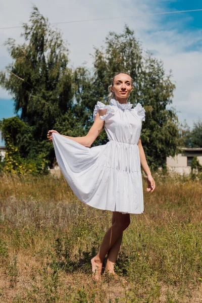 Açık renk elbiseli genç bir kız bir tarlada duruyor ve eliyle bir elbise tutuyor.