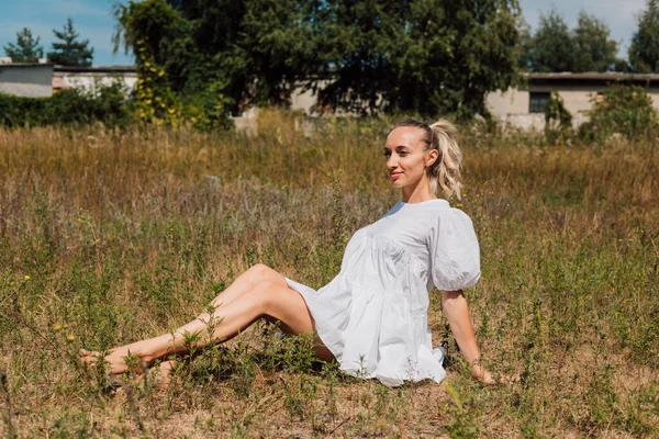 하얀 드레스를 입은 예쁜 금발의 젊은 여자가 신발 없이 잔디밭에 앉아 있다 — 스톡 사진