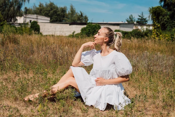 Een jonge vrouw in een witte jurk zit op het gras zonder schoenen — Stockfoto