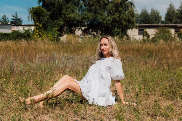 Beyaz elbiseli güzel sarışın çimlerin üzerinde ayakkabısız oturuyor. — Stok fotoğraf
