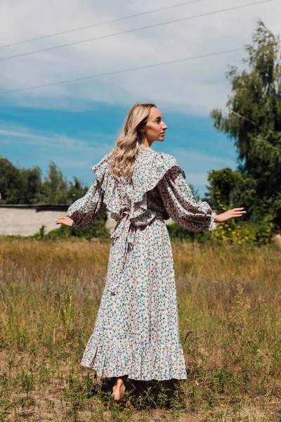 A young woman in a rustic dress walks leisurely through a field in the village — 스톡 사진