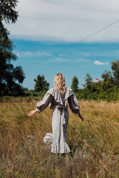 A young woman in a long country dress stands in a field with her back forward — Foto de Stock