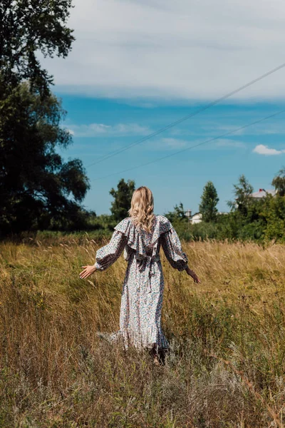A young woman in a long country dress stands in a field with her back forward — Photo