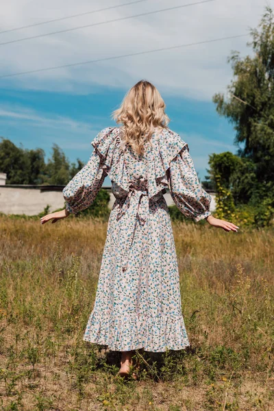 Uzun köylü elbisesi giymiş genç bir kadın sırtı önde bir tarlada duruyor.