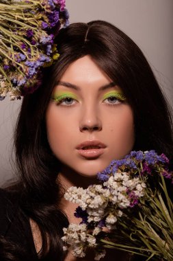 Güzel makyajlı genç bir kız mavi çiçekleri yüzüne yakın tutuyor.