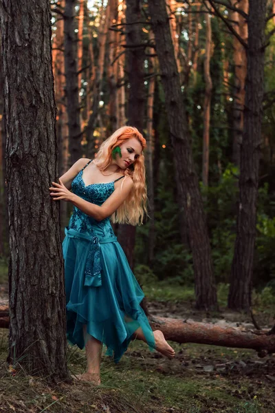 A beautiful girl with red hair stands near a tree in the forest and looks away — Foto de Stock