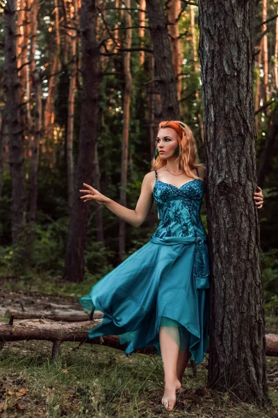 A beautiful girl with red hair stands near a tree in the forest and looks away — Foto de Stock