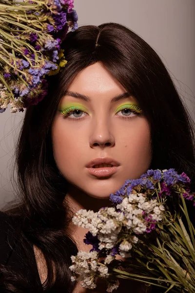 Güzel makyajlı genç bir kız mavi çiçekleri yüzüne yakın tutuyor.