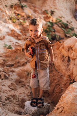 Küçük bir çocuk kumun ortasında duruyor ve elinde bir oyuncak tutuyor.