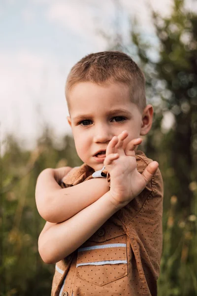 A little boy in a serious shirt stands in nature hugging himself with his hands — Stok Foto
