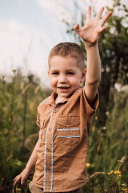 A little boy stands in a village in a field, smiling and raising his hand up.