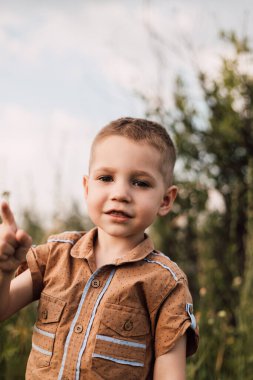 Küçük bir çocuk bir köyde duruyor, gülümsüyor ve elini kaldırıyor..