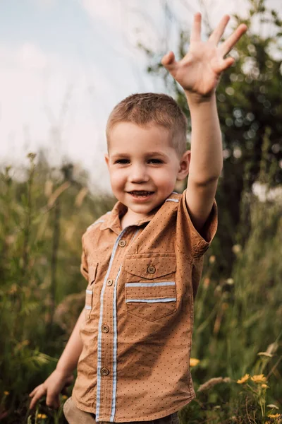 Un bambino si trova in un villaggio in un campo, sorridendo e alzando la mano. — Foto Stock