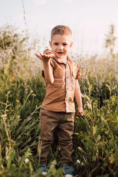 Küçük bir çocuk doğada çimlerin üzerinde duruyor ve elinde bir salyangoz tutuyor.