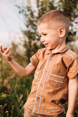 A little boy stands in a village in a field, smiling and raising his hand up.