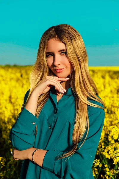 A young beautiful woman stands in a yellow rapeseed field against a blue sky — 스톡 사진