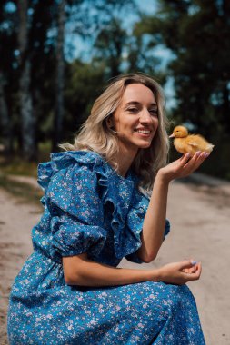 Mavi uzun elbiseli bir kız çömelmiş ve elinde bir ördek yavrusu tutuyor..