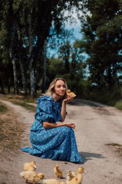 Mavi uzun elbiseli bir kız çömelmiş ve elinde bir ördek yavrusu tutuyor..