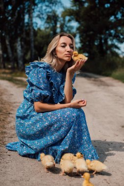 Mavi uzun elbiseli bir kız çömelmiş ve elinde bir ördek yavrusu tutuyor..