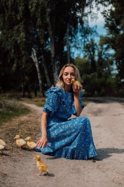 Mavi uzun elbiseli bir kız çömelmiş ve elinde bir ördek yavrusu tutuyor..