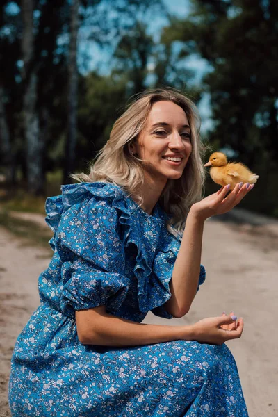 Mavi uzun elbiseli bir kız çömelmiş ve elinde bir ördek yavrusu tutuyor..