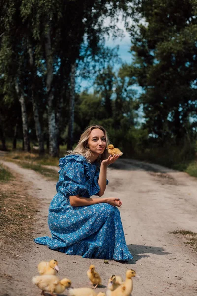Mavi uzun elbiseli bir kız çömelmiş ve elinde bir ördek yavrusu tutuyor..