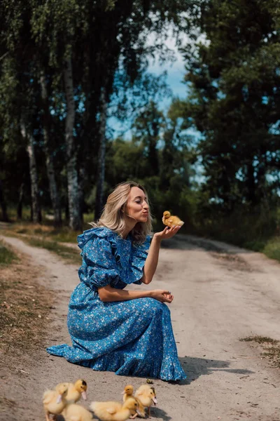 Mavi uzun elbiseli bir kız çömelmiş ve elinde bir ördek yavrusu tutuyor..