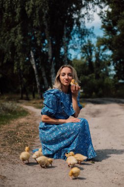 Mavi uzun elbiseli bir kız çömelmiş ve elinde bir ördek yavrusu tutuyor..
