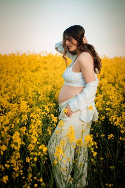 Bir eliyle saçını tutuyor. Diğer eliyle karnına sarılıyor. Sarı kolza tohumu tarlasına karşı narin bir resim..